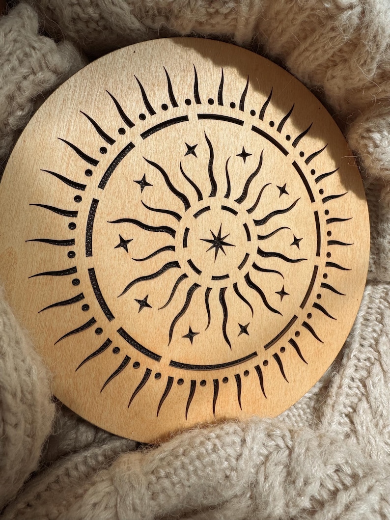 K&ouml;nnte beinhalten: Ein runder Holzuntersetzer mit einem Sonnen- und Sternenmuster. Die Sonnenstrahlen und Sterne sind ausgeschnitten, wodurch ein detailliertes Muster entsteht. Der Untersetzer ist hellbraun und liegt auf einer beigefarbenen Strickoberfl&auml;che.