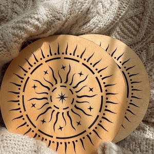 May include: Two round wooden coasters with a sunburst design. The coasters are light brown with a laser-cut pattern of a sun with rays, stars, and a central star. They rest on a textured, cream-colored knitted surface.