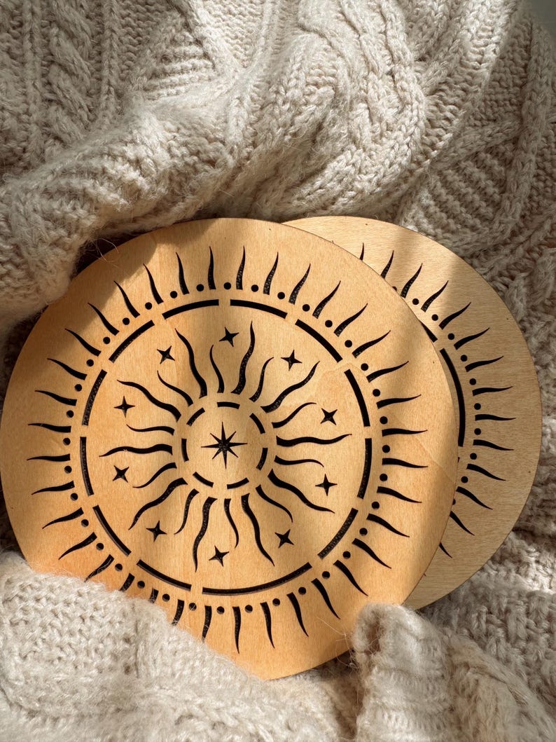 K&ouml;nnte beinhalten: Zwei runde Untersetzer aus Holz mit einem Sonnenmuster. Die Untersetzer sind hellbraun mit einem lasergeschnittenen Muster einer Sonne mit Strahlen, Sternen und einem zentralen Stern. Sie liegen auf einer strukturierten, cremefarbenen Strickoberfl&auml;che.