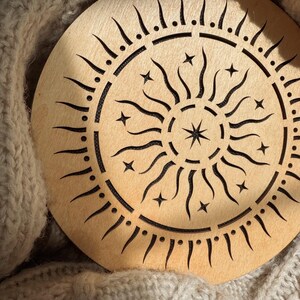 K&ouml;nnte beinhalten: Ein rundes Holzdekor mit einem Sonnenmuster. Das Sonnendesign hat strahlende Linien, Sterne und einen zentralen Stern. Das St&uuml;ck ist hellbraun und liegt auf einer beigefarbenen, gestrickten Oberfl&auml;che.