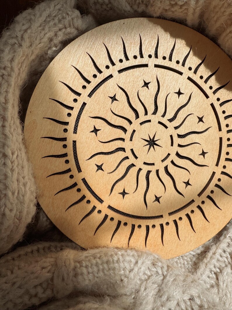 K&ouml;nnte beinhalten: Ein rundes Holzdekor mit einem Sonnenmuster. Das Sonnendesign hat strahlende Linien, Sterne und einen zentralen Stern. Das St&uuml;ck ist hellbraun und liegt auf einer beigefarbenen, gestrickten Oberfl&auml;che.