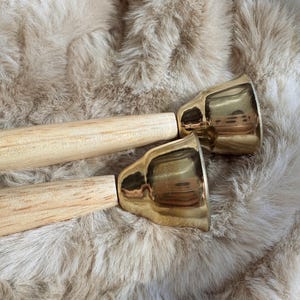 Può includere: Due campane in ottone con manici in legno poggiano su una superficie soffice e marrone chiaro. Le campane hanno una finitura dorata lucida e i manici sono in legno chiaro. Le campane sono angolate verso l'osservatore.