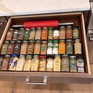May include: A kitchen drawer organized with rows of glass spice jars featuring various labels and a bright red rolling pin with silver handles positioned horizontally across the top of the spice collection within the wooden drawer frame.