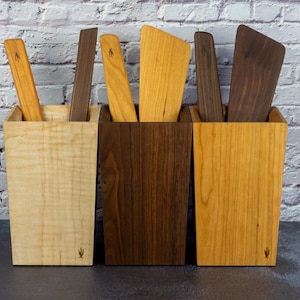 Op de afbeelding: Drie houten keukengerei houders, elk gevuld met houten spatels en lepels. De houders zijn gemaakt van licht, medium en donker hout. Elke houder heeft een klein cactus symbool op de voorkant. De achtergrond is een witte bakstenen muur.