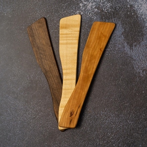 May include: Three wooden kitchen spatulas, one dark brown, one light brown with a wood grain pattern, and one light brown with a smooth finish. The spatulas are arranged in a fan shape on a gray surface.