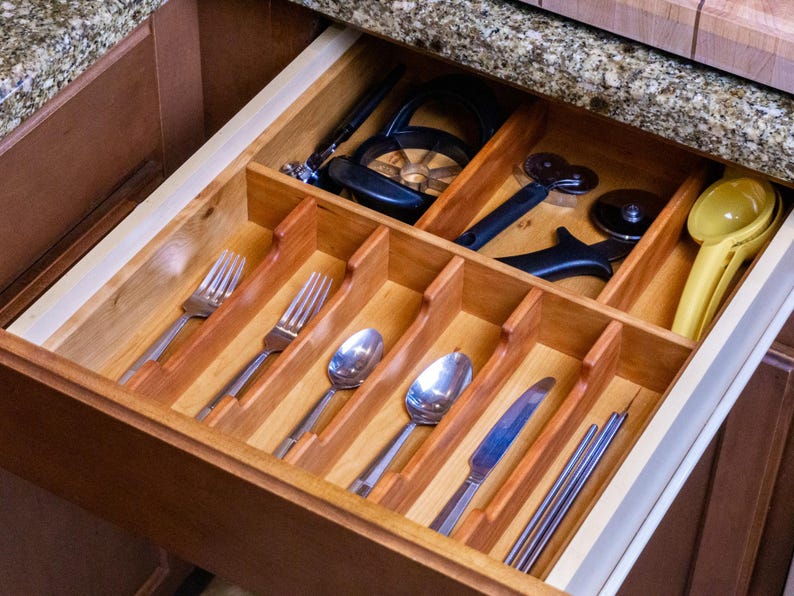 Drawer organizer made in cherry, in drawer. Neatly organized silverware set, and various cooking utensils. Granite countertops pictured.