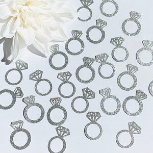 May include: Glittery silver engagement ring confetti with a large diamond-shaped gem. The confetti is scattered on a white surface, with a white flower in the background. Ideal for weddings, bridal showers, or engagement parties.