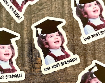 May include: A collection of stickers featuring a young child wearing a graduation cap. The stickers have a white border and the text "Look Who's Graduated". The child is wearing a white shirt and a maroon jumper.