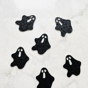 May include: Six black glitter ghost-shaped cutouts with white eye and mouth details. The Halloween-themed decorations are scattered on a white marble surface. The ghosts have a simple, cartoonish design.