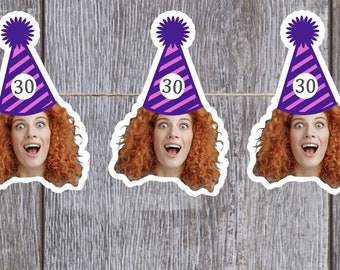 May include: Stickers featuring a woman with red curly hair wearing a purple party hat with the number 30. The stickers are hanging on a string against a gray wooden background. The woman has a surprised expression.