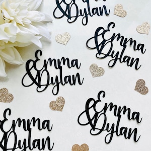 May include: Black cursive name cutouts that read "Emma & Dylan" are scattered across a white surface, along with gold glitter heart-shaped confetti. A white flower is in the upper left corner. Ideal for wedding decor.