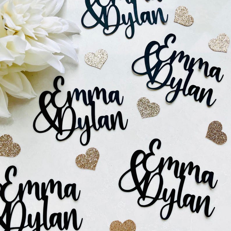 May include: Black cursive name cutouts that read "Emma & Dylan" are scattered across a white surface, along with gold glitter heart-shaped confetti. A white flower is in the upper left corner. Ideal for wedding decor.