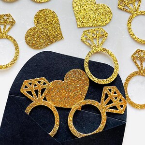 May include: Gold glitter engagement ring and heart-shaped decorations. The ring decorations have a diamond-shaped top. Some of the decorations are emerging from a black envelope. The background is white.