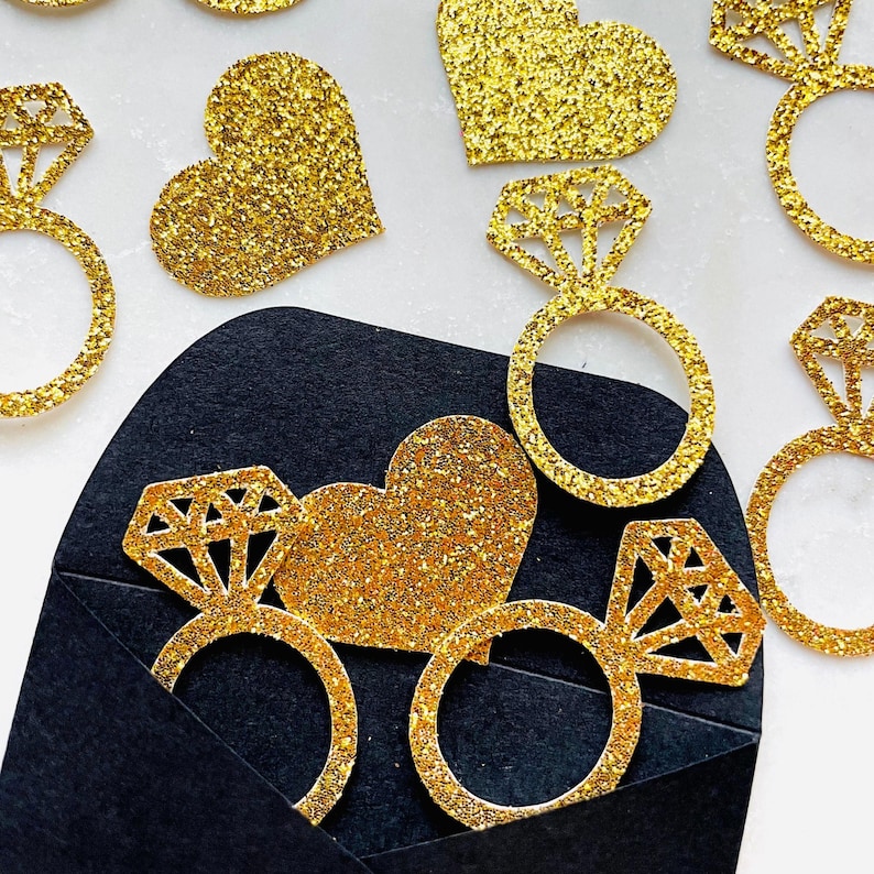 May include: Gold glitter engagement ring and heart-shaped decorations. The ring decorations have a diamond-shaped top. Some of the decorations are emerging from a black envelope. The background is white.
