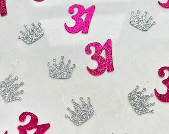 Könnte beinhalten: Streukonfetti mit der Zahl "31" in pinkfarbenem Glitzer und silbernen Glitzerkronenformen, verstreut auf einer weißen Oberfläche. Geeignet für eine Geburtstagsfeier.