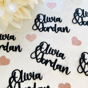 Può includere: Ritagli neri in corsivo dei nomi "Olivia & Jordan" sono disposti su una superficie bianca, intervallati da piccoli cuori glitterati color oro rosa. Un grande fiore bianco sporco si trova nell'angolo in alto a sinistra. Ideale per matrimoni.