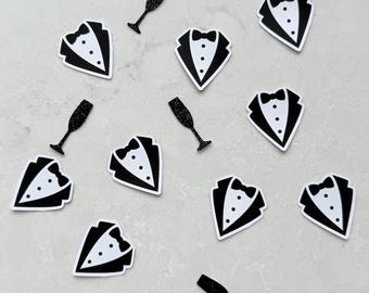 May include: Scattered black and white tuxedo-shaped stickers with bow ties and button details. Also, scattered black glitter champagne flutes. The stickers are on a light gray surface.