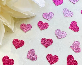 Könnte beinhalten: Auf einer hellen Oberfläche sind zahlreiche herzförmige Glitzerverzierungen in verschiedenen Rosa- und Fuchsia-Tönen verstreut. Eine große weiße Blume ist teilweise in der oberen linken Ecke zu sehen. Diese dekorativen Herzen sind ideal für Bastelarbeiten und Valentinstag.