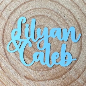 May include: A light blue, laser-cut wooden sign with the names "Lilyan & Caleb" in a cursive font. The sign is placed on a wooden surface with visible wood grain.