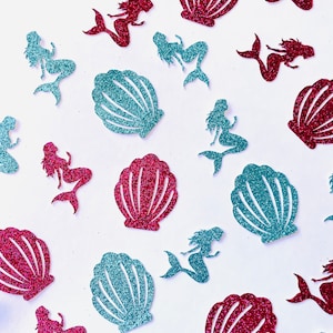 Op de afbeelding: Een patroon van glitterende zeemeermin- en schelpenuitsparingen in tinten teal en magenta op een witte achtergrond. De zeemeerminfiguren staan in verschillende poses en de schelpen zijn waaiervormig.