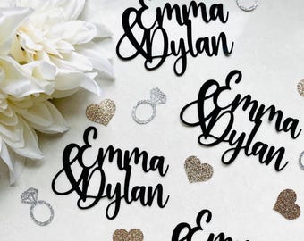 May include: Black cursive cutouts of "Emma & Dylan" are scattered among glitter hearts and diamond ring decorations. A large white flower is in the upper left corner. The decorations are likely for a wedding or engagement celebration.