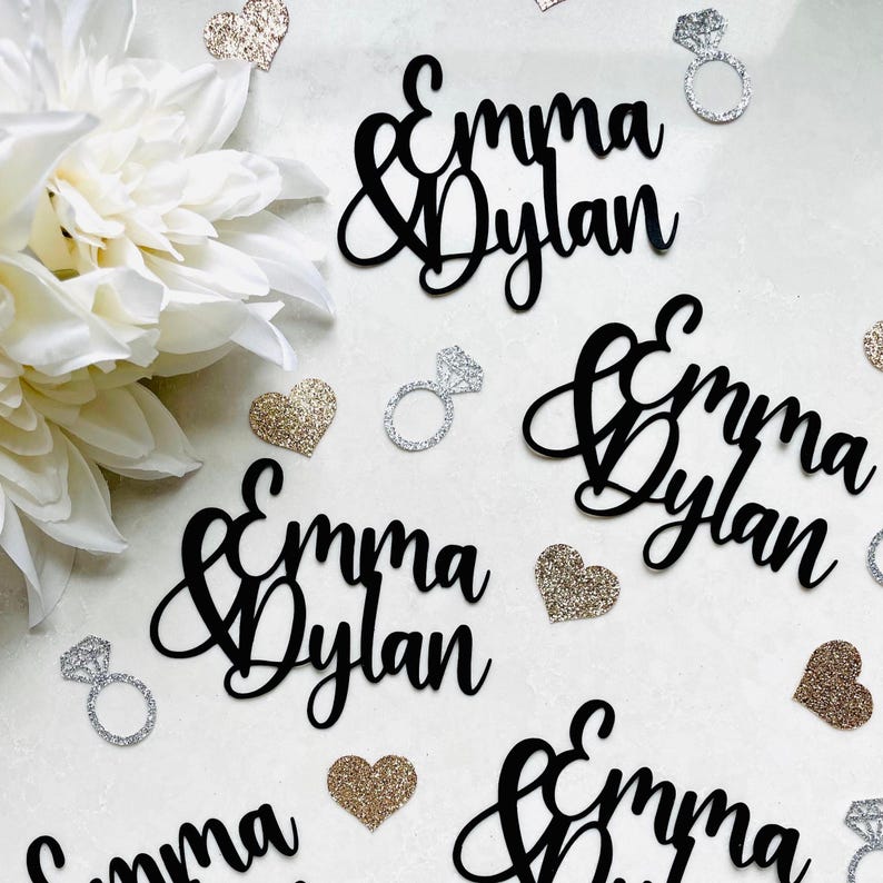 May include: Black cursive cutouts of "Emma & Dylan" are scattered among glitter hearts and diamond ring decorations. A large white flower is in the upper left corner. The decorations are likely for a wedding or engagement celebration.