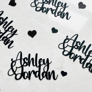 May include: Black die-cut name cutouts that read "Ashley Jordan" in a cursive font, scattered across a white surface. Several small black glitter hearts are also scattered around the names. The names are likely for a wedding or event.