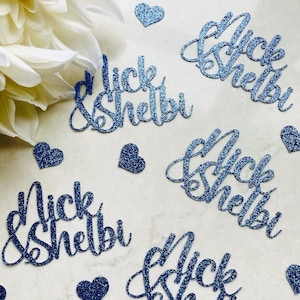 May include: Blue glitter cutouts spelling "Nick & Shelbi" in a cursive font, scattered with small glitter hearts. A white flower is in the upper left corner. Ideal for wedding or party decorations.