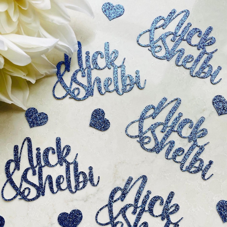May include: Blue glitter cutouts spelling "Nick & Shelbi" in a cursive font, scattered with small glitter hearts. A white flower is in the upper left corner. Ideal for wedding or party decorations.
