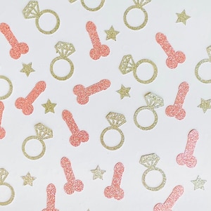May include: Glittery gold engagement rings, pink phalluses, and gold stars are scattered across a white surface. The shapes are likely confetti, perfect for a bachelorette party or bridal shower. The shapes are all glittery.