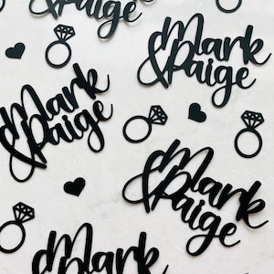 May include: Black paper cutouts of the names "Mark & Paige", diamond rings, and hearts. The names are in a cursive font. The diamond rings have a detailed diamond shape. The hearts are solid black.
