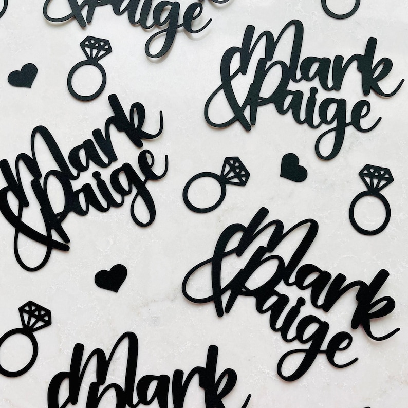 May include: Black paper cutouts of the names "Mark & Paige", diamond rings, and hearts. The names are in a cursive font. The diamond rings have a detailed diamond shape. The hearts are solid black.