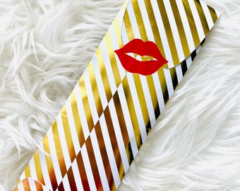Könnte beinhalten: Eine gold-weiß gestreifte Geschenkverpackung mit einem roten Lippen-Emblem. Der Umschlag ist geschlossen und hat eine Klappe. Der Hintergrund ist eine weiße, flauschige Oberfläche.