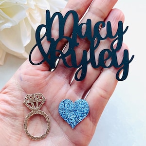 May include: A hand holds three decorative items: a black cursive cutout with the names "Macy & Ryley", a gold glitter ring, and a blue glitter heart. The ring features a diamond-shaped top.
