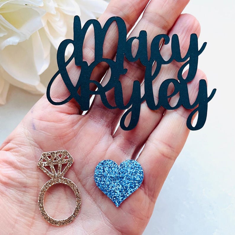 May include: A hand holds three decorative items: a black cursive cutout with the names "Macy & Ryley", a gold glitter ring, and a blue glitter heart. The ring features a diamond-shaped top.