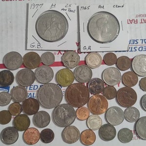May include: A collection of various coins, including two encased coins with the years 1977 and 1965. The coins are of different sizes and colors, scattered on a white surface with red stripes. Some coins are in protective sleeves.