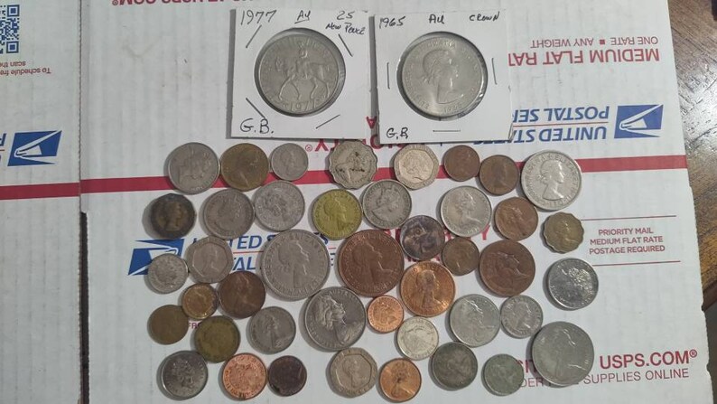 May include: A collection of various coins, including two encased coins with the years 1977 and 1965. The coins are of different sizes and colors, scattered on a white surface with red stripes. Some coins are in protective sleeves.