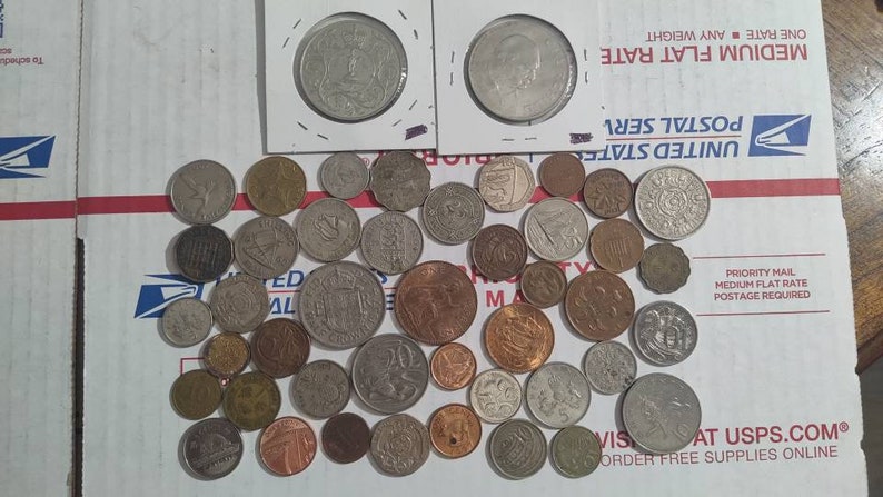 May include: A collection of various coins, including silver and copper, scattered on a white surface. Two coins are encased in protective sleeves. The coins vary in size and design, showcasing historical currency.