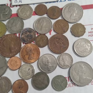 May include: A collection of various coins, including silver and copper examples, some featuring profiles of historical figures. One coin is in a protective sleeve with the text "1965 Au Crown". The coins are scattered on a white surface.