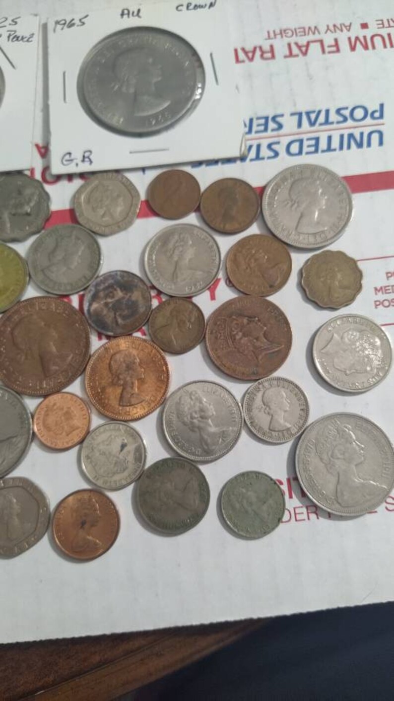 May include: A collection of various coins, including silver and copper examples, some featuring profiles of historical figures. One coin is in a protective sleeve with the text "1965 Au Crown". The coins are scattered on a white surface.