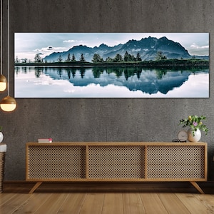 May include: A panoramic photograph of a mountain range reflected in a still lake. The mountains are a dark blue color and the lake is a light blue color. The sky is a light blue color with some clouds. There are trees along the shore of the lake.