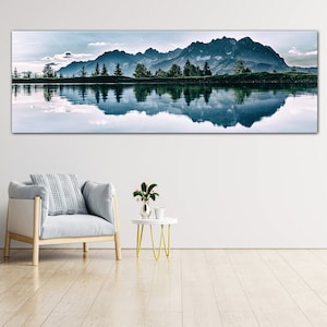 May include: A panoramic photograph of a mountain range reflected in a still lake. The mountains are a dark blue color and the lake is a light blue color. The sky is a light blue color with white clouds. There are trees along the shore of the lake.