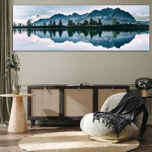 May include: A panoramic photograph of a mountain range reflected in a still lake. The mountains are a dark blue-grey color and the lake is a light blue. The sky is a light blue with some white clouds. The image is framed and hanging on a wall above a wooden cabinet and a light grey armchair with a black and white throw blanket.
