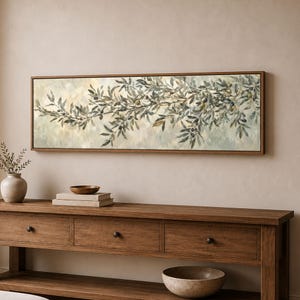 May include: A rectangular wall art piece featuring a horizontal painting of olive branches with dark green leaves and small olives. The artwork is enclosed in a thin brown wooden frame and displays a soft, muted background with blended tones of cream and light green.