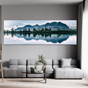 May include: A panoramic photograph of a mountain range reflected in a still lake. The mountains are a dark blue color and the lake is a light blue color. The sky is a light blue color with white clouds. The image is a long horizontal format.