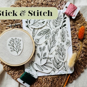 May include: A set of white fabric stickers with black line drawings of various floral designs. The stickers are arranged on a white sheet of paper. The text "Stick & Stitch" is written in green on a light green background. There are two spools of embroidery floss, one green and one pink, and a small wooden embroidery hoop with a floral design embroidered on it.