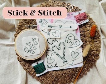 May include: A craft kit with the text "Stick & Stitch" includes an embroidery hoop with a floral design, various fabric patches with heart and arrow designs, and spools of pink and green thread. The kit is arranged on a woven mat.