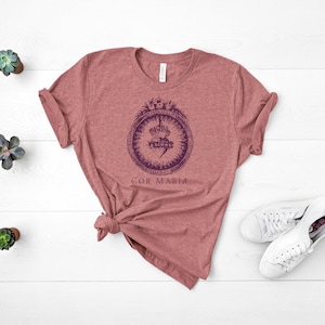 May include: A heather red t-shirt with a vintage-style illustration of a heart with a crown and thorns. The text "COR MARIA" is printed below the illustration.