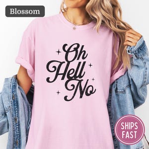 Könnte beinhalten: Ein hellrosa T-Shirt mit kurzen Ärmeln und dem schwarzen Schriftzug Oh Hell No in einer stilisierten Schreibschrift, akzentuiert durch kleine vierzackige Sternmotive. Das T-Shirt hat einen Rundhalsausschnitt und eine blaue Jeansjacke.