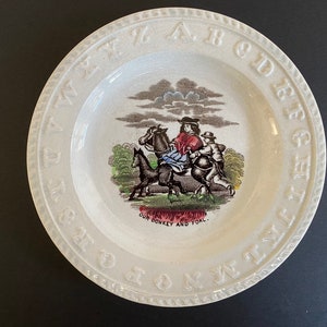 May include: Cream-colored ceramic plate with a raised alphabet border. The center features a colorful illustration of a person riding a horse, with the text "OUR DONKEY AND FOAL." A vintage decorative piece.